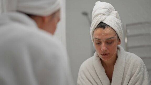 女士使用保湿泡沫洗脸和微笑早上的护肤程序年轻的女学生头上缠着毛巾在水疗沙龙照镜子准备化妆