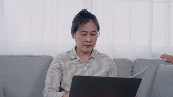 4K亚洲女性在客厅的沙发上用笔记本电脑工作手里拿着纸她的儿子蒙着眼睛从后面跑了过来高兴地逗着妈妈他们相视一笑