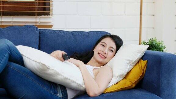 年轻的亚洲女子一边拿着遥控器一边躺在家里客厅的沙发上看电视人们享受着生活方式