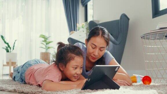 亚洲蹒跚学步的小女孩女儿和妈妈在地毯上看数码平板电脑有乐趣在家里的客厅快乐的时刻