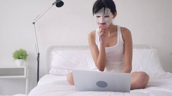 美丽年轻的亚洲女性带着单面膜坐在卧室的床上在笔记本电脑上工作美丽女孩在家里用面膜护理皱纹皮肤护理健康和健康