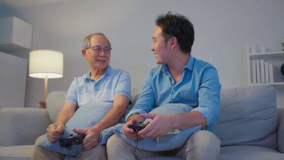 亚洲快乐家庭晚上在客厅玩游戏机游戏年长的老人年长的父亲和年轻英俊的男性坐在沙发上感到兴奋和享受花时间玩操纵杆游戏在家里