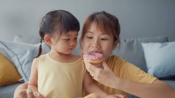 快乐快乐的亚洲家庭妈妈和蹒跚学步的女孩吃着甜甜圈在家里客厅的沙发上放松享受