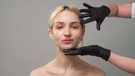 戴着黑色手套的整形外科医生的手触摸着一个女人的脸在浅色的背景下一个美丽的女孩没有化妆皮肤干净外科手术和美容面部注射
