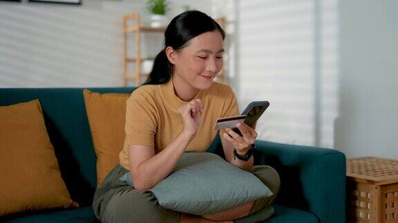 亚洲女人开心地微笑着坐在沙发上用智能手机和信用卡在家里网上购物