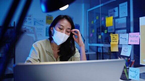 快乐亚洲女商人戴着医用口罩在新常态下进行社交距离以预防病毒同时在晚上工作时使用笔记本电脑