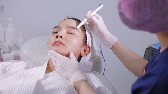 亚洲年轻漂亮的女性穿着浴袍和毛巾缠头巾躺在医生面前在前额皮肤上做真皮皮肤针刺治疗亚洲年轻漂亮女性在脸上做微针按摩使脸部绷紧、提升