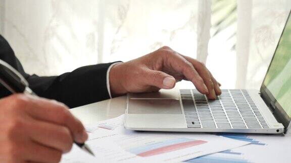 特写商人双手工作使用笔记本电脑财务业务文件阅读报告文书工作电子表格在办公桌上男人用手打键盘在办公笔记上写查财务图表