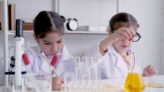 4K两个小女孩坐在一起在科学教室做实验小女孩用试管里的水做实验她的朋友是一个记录所有实验的人