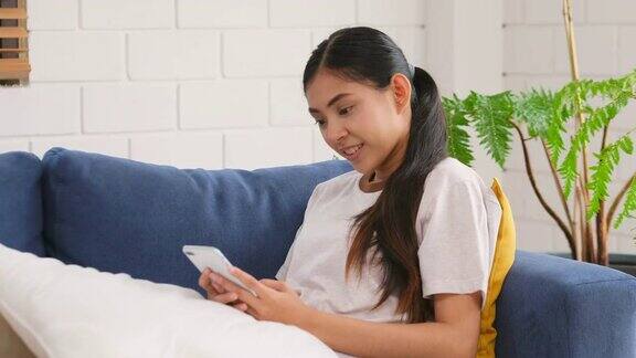 年轻美丽的亚洲女人使用智能手机与幸福而坐在家里的沙发上人们在电话生活方式