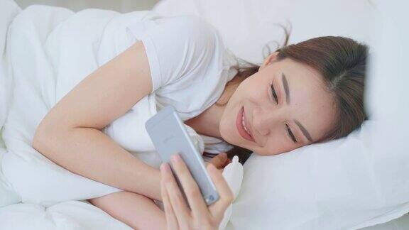 亚洲美女早上在家里的床上用手机聊天快乐的休闲美女躺在卧室里享受假期周末在家里用智能手机和科技交流