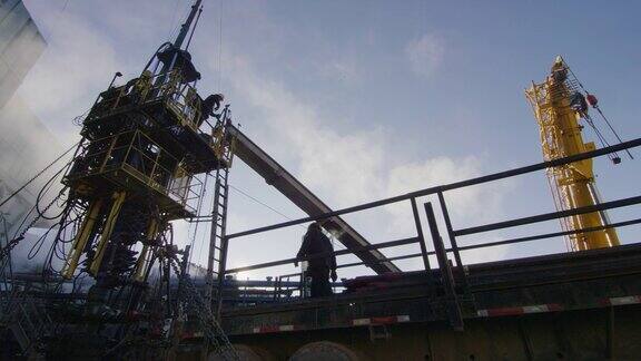 在一个寒冷的冬天的早晨油田工人在石油和天然气钻井平台的一个高高的金属平台上架设钻杆的慢镜头