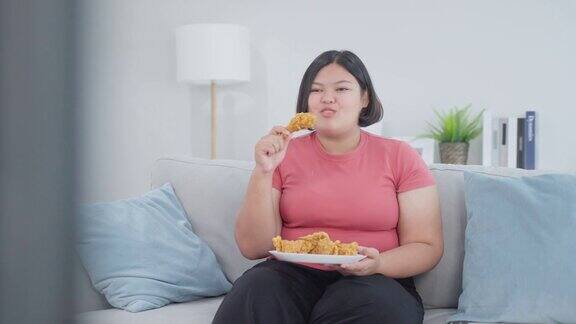 亚洲胖女人看电视的时候吃不健康的食物年轻漂亮的饿胖女孩感到快乐享受在家里客厅的沙发上看电视节目和咬炸鸡