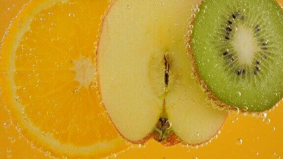 将成熟的橙子、苹果和猕猴桃切片放入透明的水中在橙色背景上形成气泡