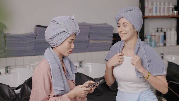 两个漂亮的女孩用灰色浴衣裹着头发坐在洗头区一边聊天一边从手机上挑选发型发廊工作室秉承发型时尚、美容、时尚的理念
