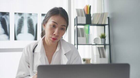 年轻的亚洲女医生穿着白色医疗制服带着听诊器使用电脑笔记本电脑在健康诊所或医院的办公桌上与病人进行视频电话会议