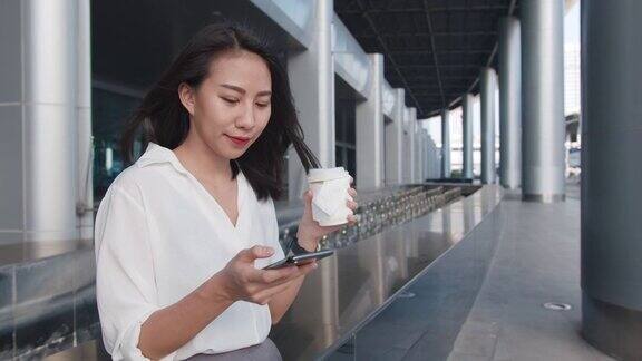 亚洲女商人穿着时尚的办公室服装拿着一次性纸杯热饮站在城市现代城市的户外使用智能手机