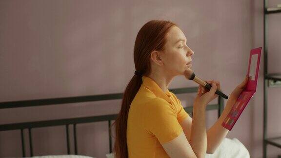 天生丽质的红发年轻女子用化妆刷化妆在家里的卧室里敷粉