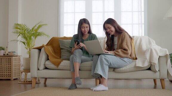 两个亚洲年轻女子开心地微笑着在家里客厅的沙发上使用笔记本电脑