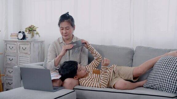 亚洲单身母亲坐在沙发上用笔记本电脑工作男孩困了把平板电脑递给母亲然后躺在他的腿上睡着了