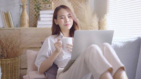 年轻的亚洲妇女使用笔记本电脑在沙发客厅在家里舒适快乐的工作在家里