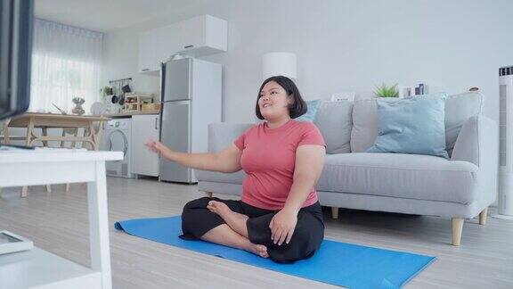 亚洲超大的女人在客厅里做瑜伽锻炼超重的运动女孩坐在自由体操伸展手根据在线教练的指导为健康减肥