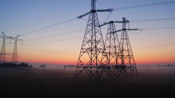 鸟瞰图高电压钢电力塔在田野覆盖着雾乡村的雾蒙蒙的清晨黎明无人机在输电线路上空低空飞行电塔线日出日落