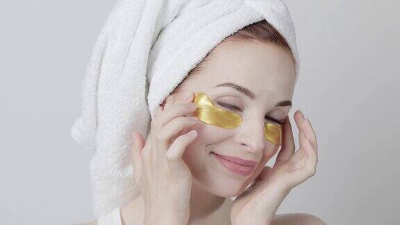 美丽微笑的女人头上的毛巾在家庭水疗过程中应用金色的眼罩