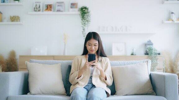 快乐的年轻亚洲女人在家里舒适的沙发上放松地用智能手机发短信微笑的女孩用手机聊天用小工具浏览无线互联网在家网上购物