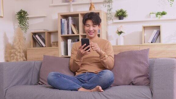 一名亚洲男子坐在家里的沙发上玩智能手机男子一边工作一边使用社交媒体应用在家工作