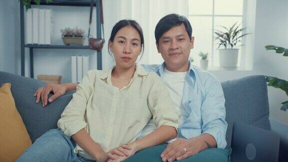 年轻的亚洲夫妇与休闲坐在沙发上看相机在线呼叫挥手手势与家人和朋友在家里的客厅交谈