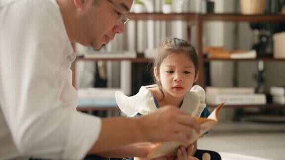 一位年轻的父亲正和女儿坐在客厅的地板上给她读一本书