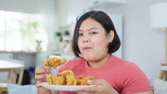 亚洲胖女人的肖像在家里吃不健康的食物漂亮的大码女孩感到饥饿和快乐在客厅的沙发上咬炸鸡微笑看着摄像机