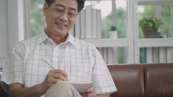 一位上了年纪的亚洲人坐在客厅里带着快乐的微笑在家里工作的科技理念