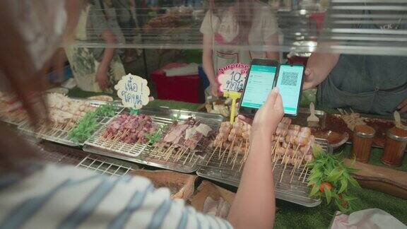 一名亚洲女子在夜市买了猪肉串、肉串和蔬菜串后用智能手机扫描条形码付款泰国的街头小吃