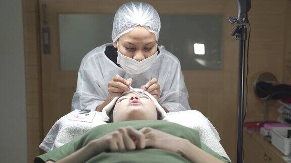 亚洲化妆师制作睫毛沙龙程序女人的睫毛扩展专业人员佩戴医用口罩帽和个人防护服在新冠肺炎疫情期间提供卫生服务年轻健康的皮肤