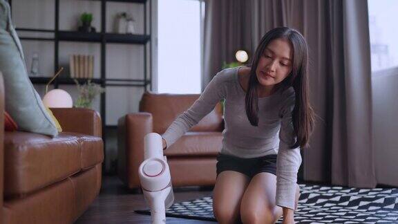 穿着t恤和短裤的亚洲年轻美女正在家里明亮舒适的房间里用吸尘器清洁地毯她用的是现代无绳吸尘器电器和有趣