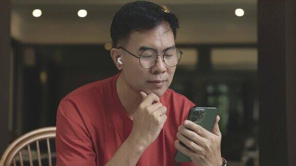 戴着眼镜的亚洲中年男子正在用智能手机和蓝牙耳机轻松愉快地观看娱乐电影通过手机输入通信信息