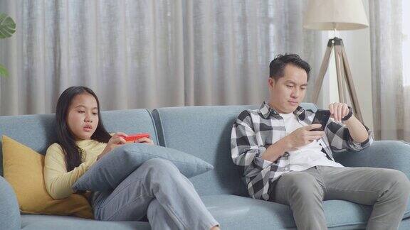 亚洲父亲和女儿坐在家里客厅的沙发上一边用智能手机刷社交媒体