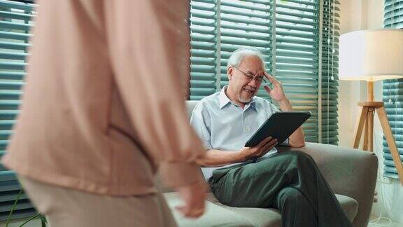 一名退休的亚洲老人坐在沙发上用平板电脑看网络新闻时感到头痛