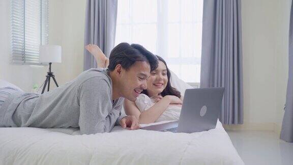 亚洲年轻夫妇躺在床上用笔记本电脑在卧室看电影漂亮的新婚男女穿着睡衣感到幸福和放松清晨醒来后看电脑上的节目