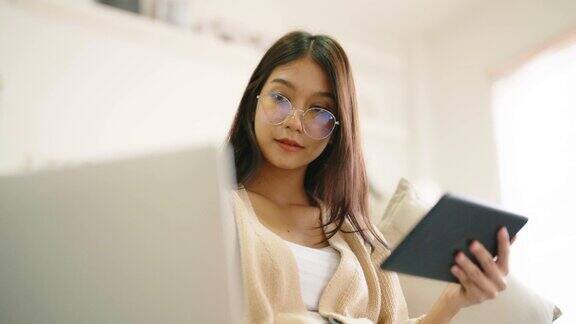 年轻的亚洲妇女戴着眼镜坐在家里的沙发上使用平板电脑