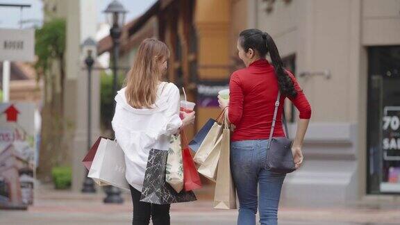 后视图两个女士走在人行道上在购物中心购物后回家的路上互相交谈他们的手拿着购物袋和冷饮
