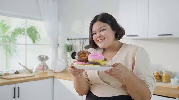 亚洲特大号女人的肖像在厨房的盘子里拿着甜甜圈年轻迷人漂亮的大码女孩感到幸福享受吃甜蜜的不健康食品和微笑看着摄像机在家里