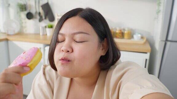 亚洲超大视频博主胖女人在家厨房吃不健康食品年轻迷人的漂亮的大码女孩拿着相机和记录视频直播咬甜甜圈在家里的桌子上
