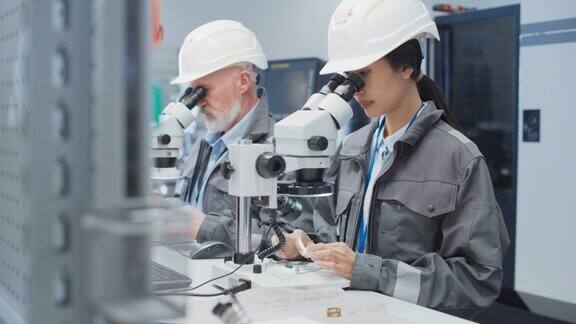 电子制造工厂:两个工程师通过显微镜检查小型工业设备部件技术人员测试新的工厂部件以保证生产质量