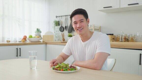 亚洲活跃帅哥拒绝甜甜圈喜欢吃健康食品年轻有魅力的男性在厨房的碗里吃蔬菜沙拉在桌子上喝干净的水感觉很开心饮食保健理念