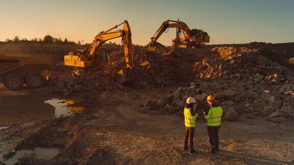 空中无人机拍摄的建筑工地在阳光明媚的晚上:工业挖掘机挖掘岩石并把它们装上卡车土木工程师和检查员观察过程讨论房地产项目