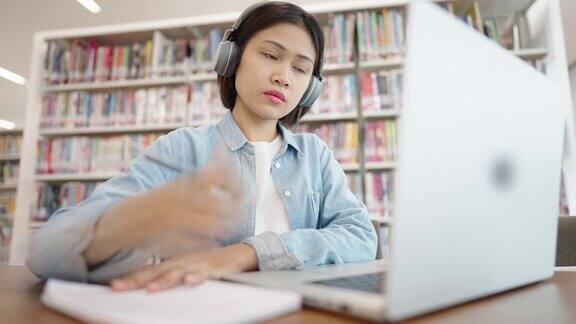 亚洲女大学生在图书馆戴着耳机在笔记本电脑上上网学习在图书馆为考试而学习