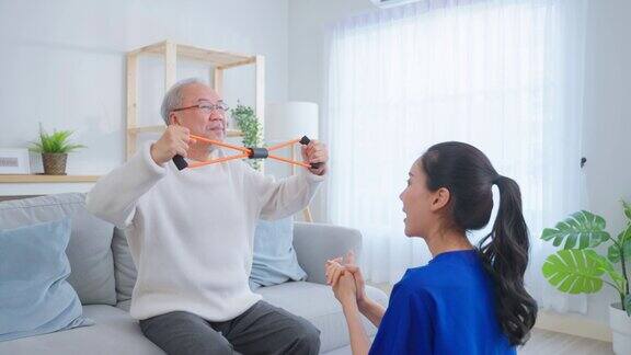 亚洲老年男子在护士的支持下做物理治疗老年男性患者坐在客厅的沙发上在养老院的治疗师医生的指导下进行哑铃锻炼